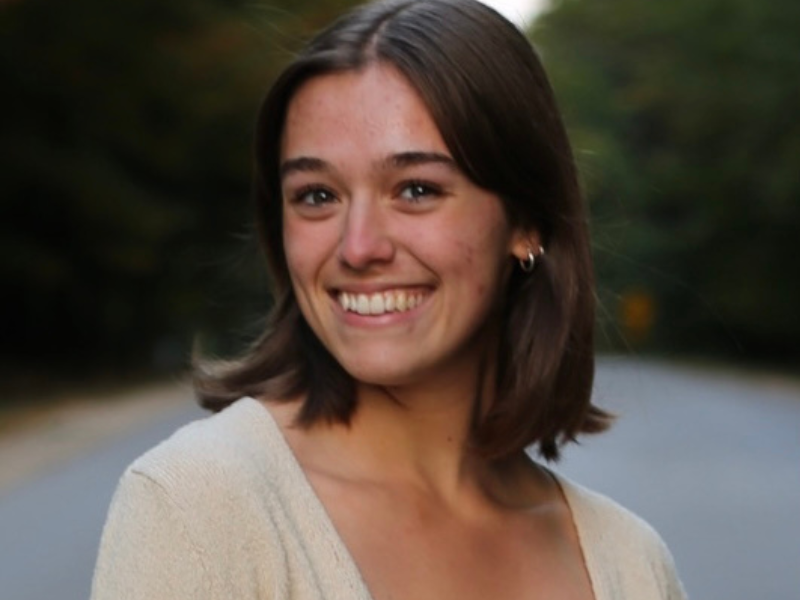A young woman with shoulder-length brown hair smiles warmly at the camera, wearing a light sweater, with a blurred tree-lined road in the background.