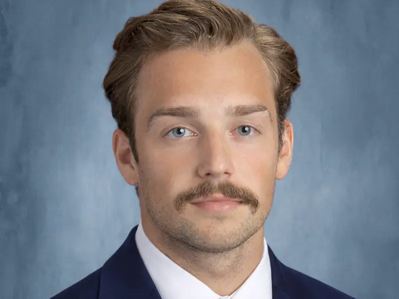 A young man with short, styled hair and a mustache, wearing a dark suit and white shirt, against a soft blue background.