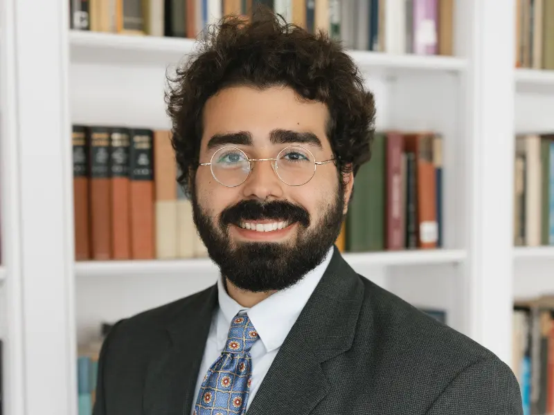 A smiling man with curly hair and glasses, dressed in a suit and tie, stands in front of a bookshelf filled with books.
